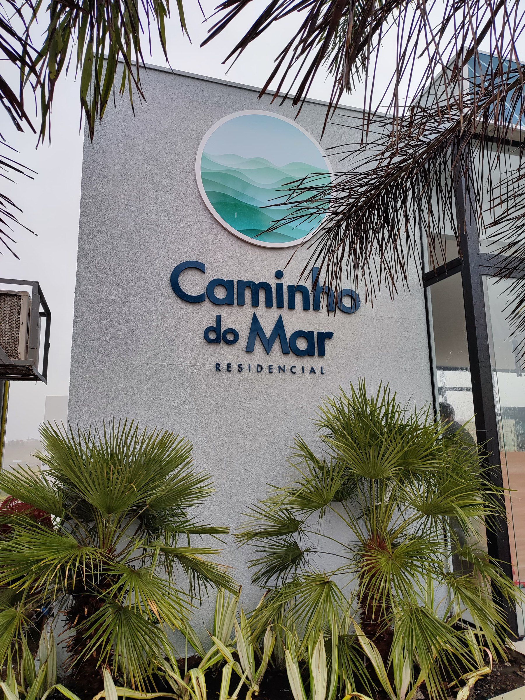Letreiro Caminho do Mar Residencial em letras caixa na parede do estande, entre palmeiras
