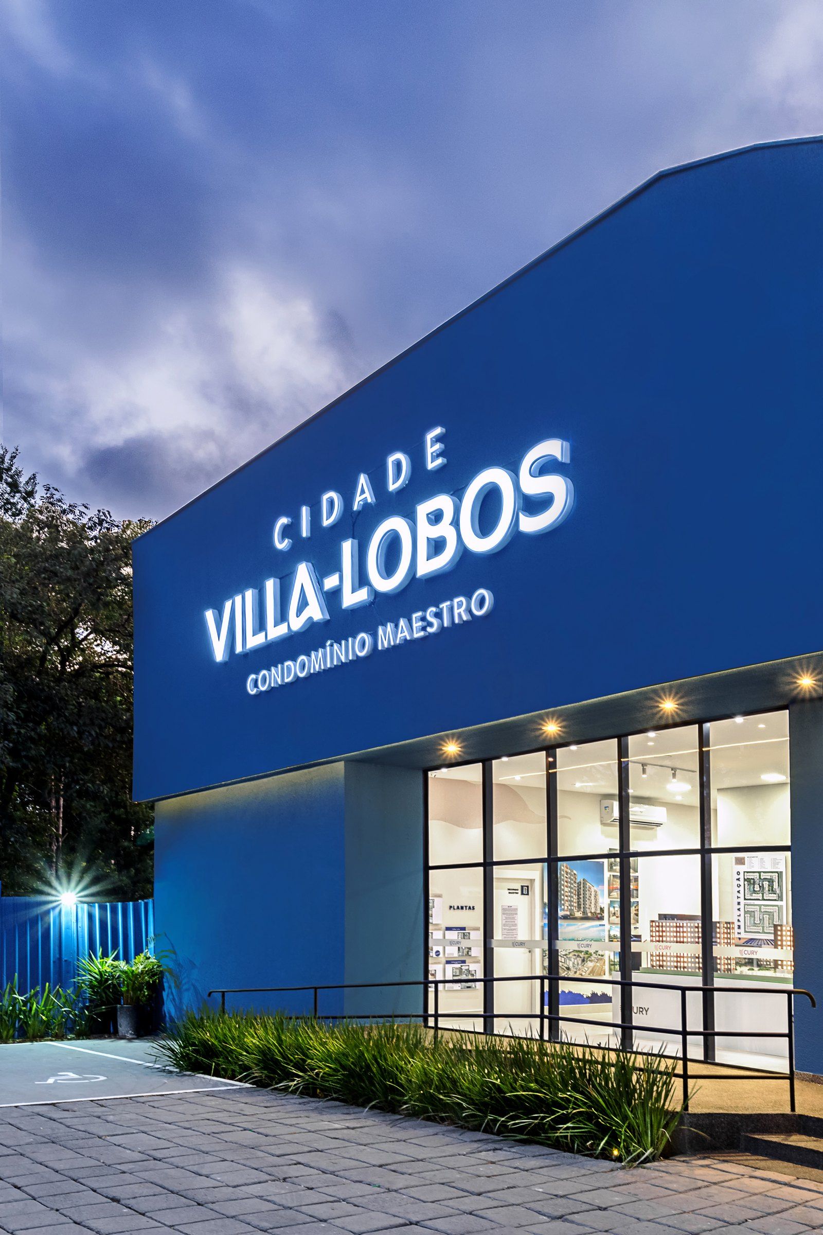 Fachada azul curva do estande Cidade Villa-Lobos, da Cury, com letreiro iluminado ao anoitecer