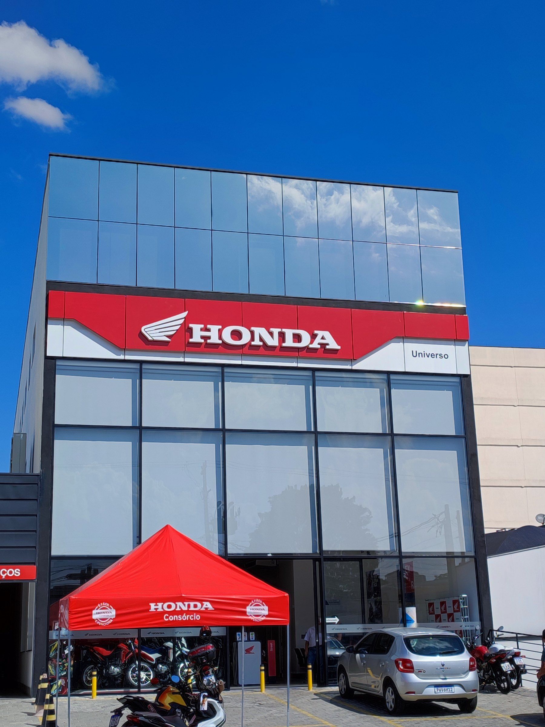 Concessionária Honda com fachada de vidro e faixa vermelha com o letreiro Honda em letras caixa, sob céu azul