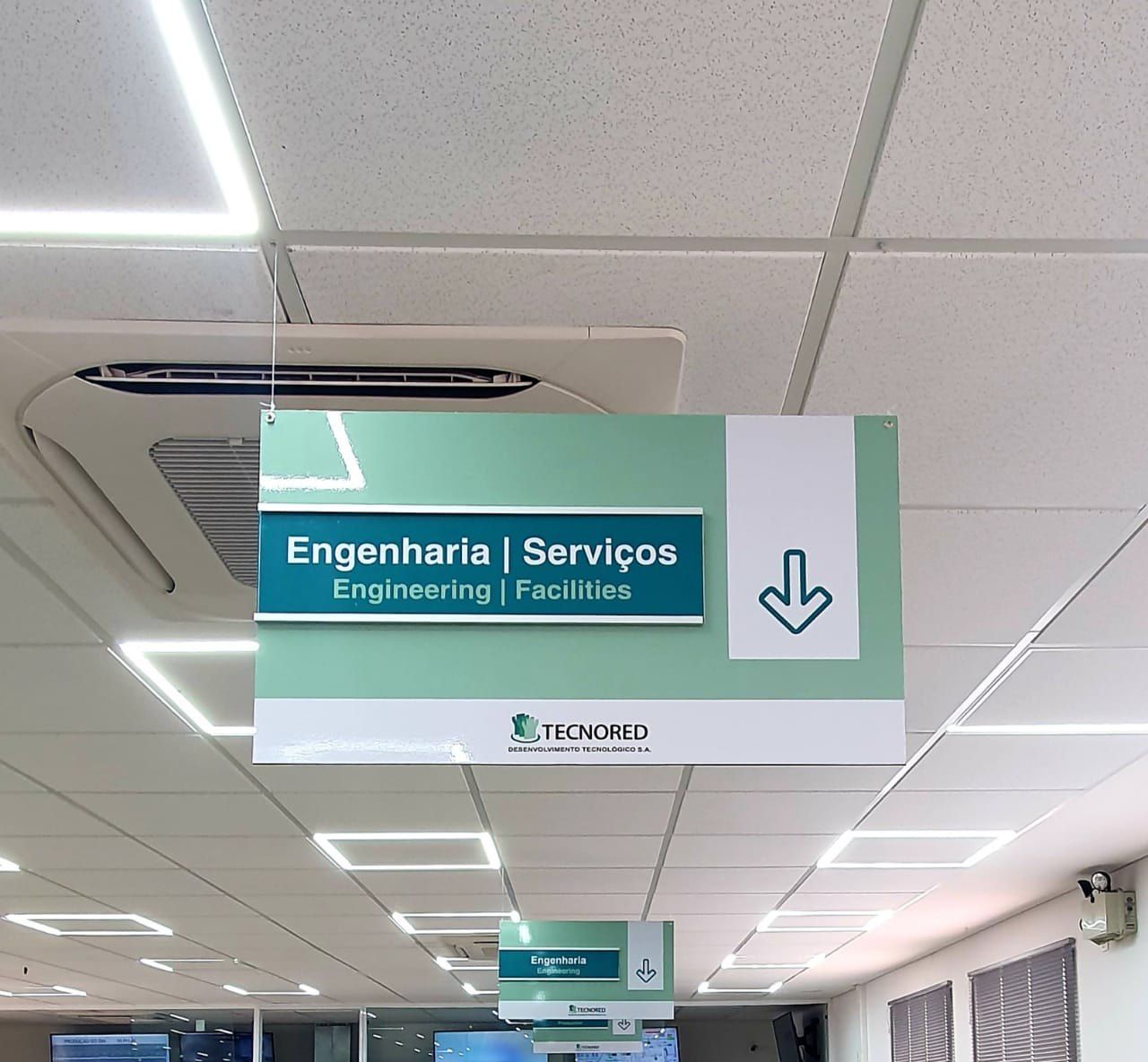 Placa suspensa Engenharia e Serviços, em português e inglês, no teto do escritório da Tecnored